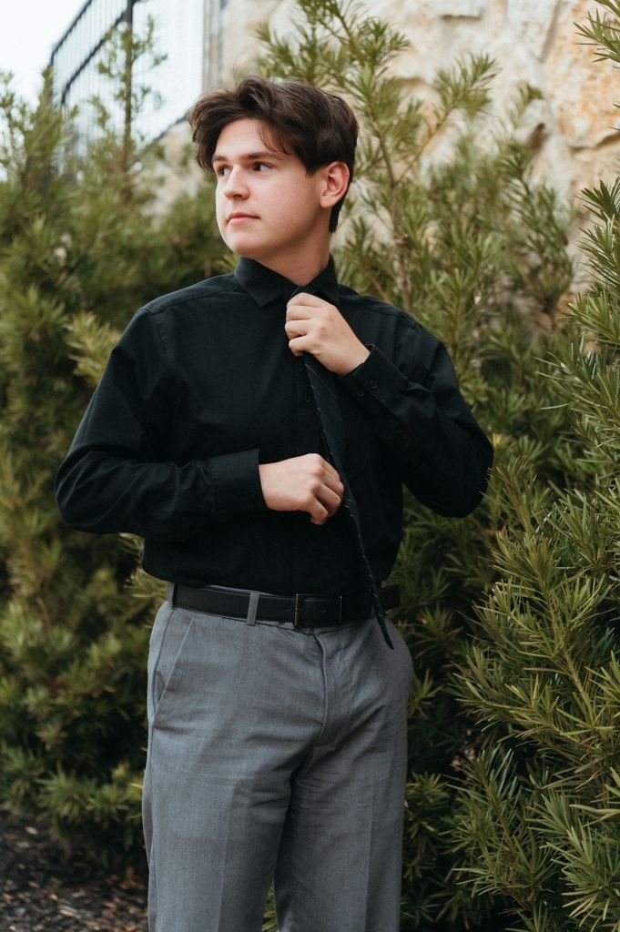 Young man in black dress shirt and gray trousers adjusting collar outdoors near green shrubs.