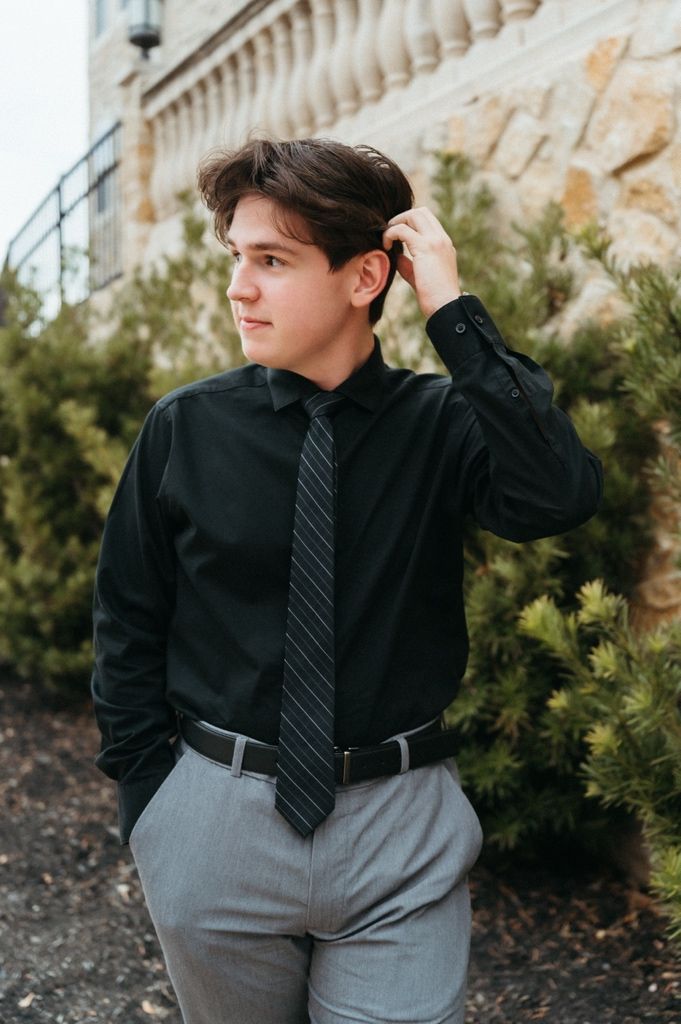 Young man in black dress shirt and striped tie with gray trousers poses outdoors near evergreen shrubs.