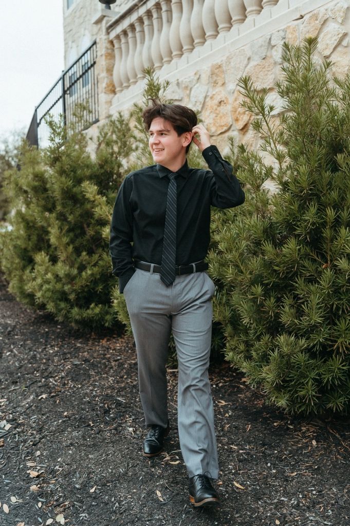 Young man in black dress shirt, striped tie, and gray trousers posing outdoors near evergreen shrubs and stone wall.