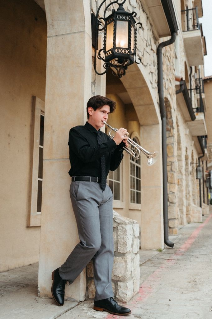 Young male trumpet player in black shirt and gray pants performing outdoors against a stone building with vintage lantern.