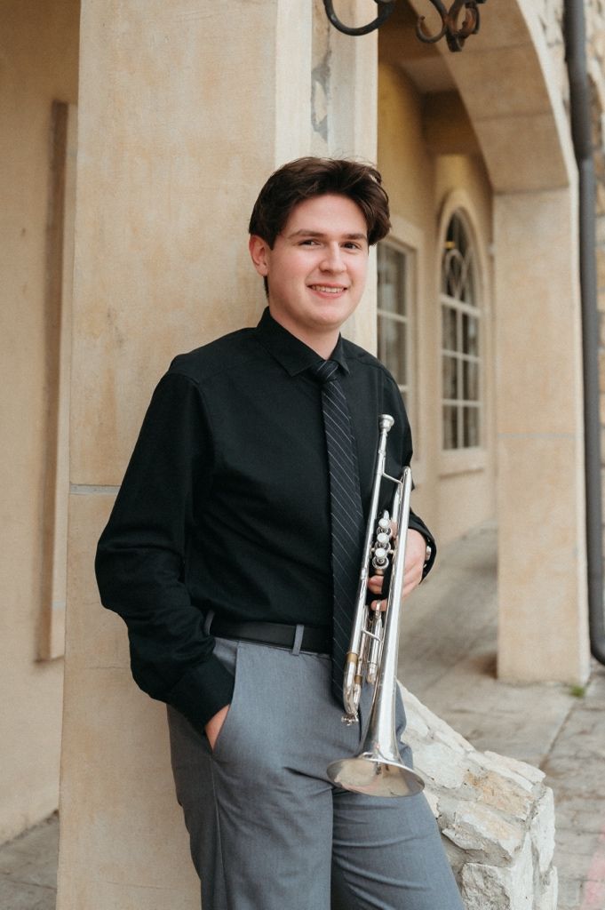 Young male trumpet player in black shirt and gray pants poses with silver trumpet against stone building exterior.