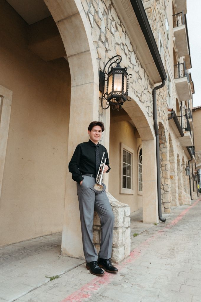 Young musician in black shirt and gray pants holds trumpet while leaning against stone archway building outdoors.