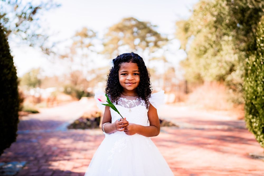 Young Black girl in white flower girl dress holding a tulip, smiling outdoors on a red brick path.