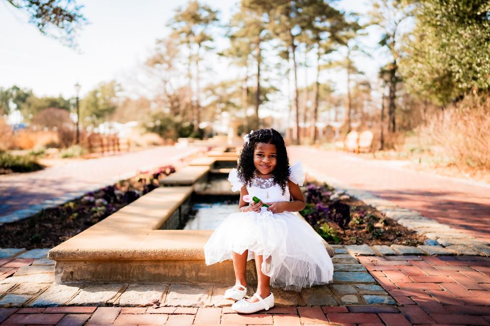 Young girl in white tutu dress sitting on brick pathway in a garden, smiling joyfully on a sunny day.
