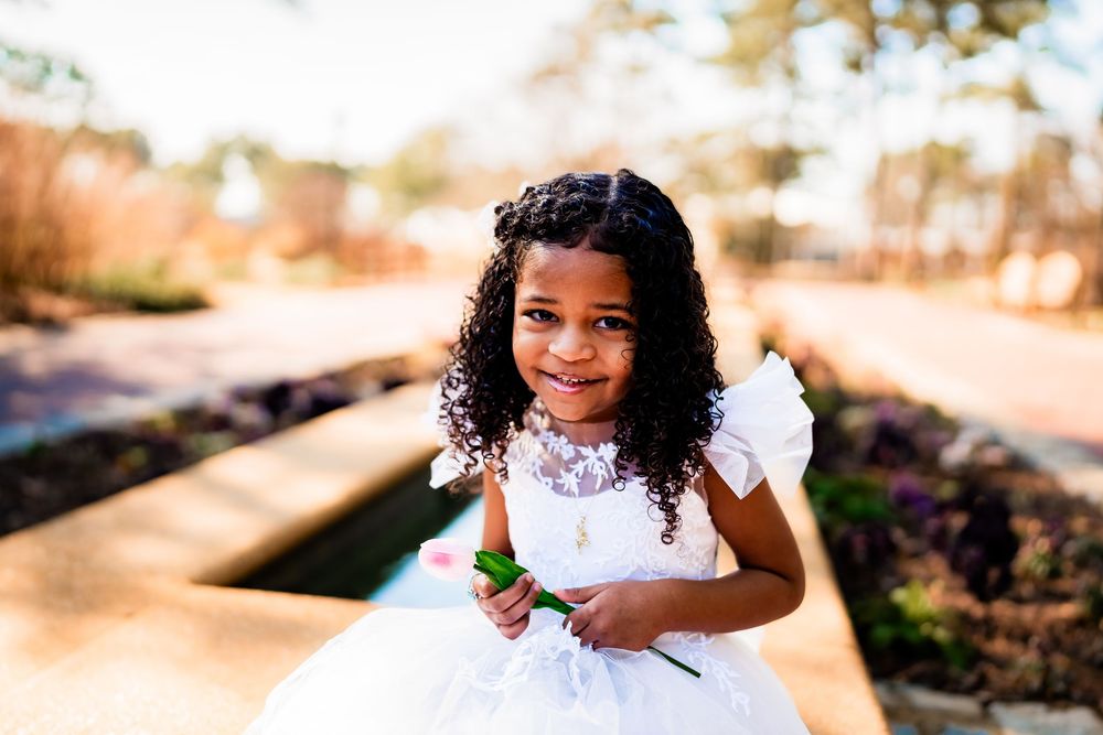 A young Black girl in a white tulle dress smiles outdoors, holding a flower in a sunny garden setting.