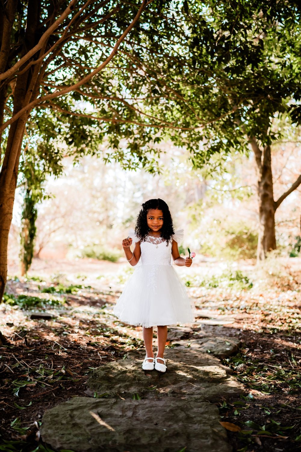 Young girl in white tulle dress standing on stone path surrounded by lush green trees in sunlit garden.