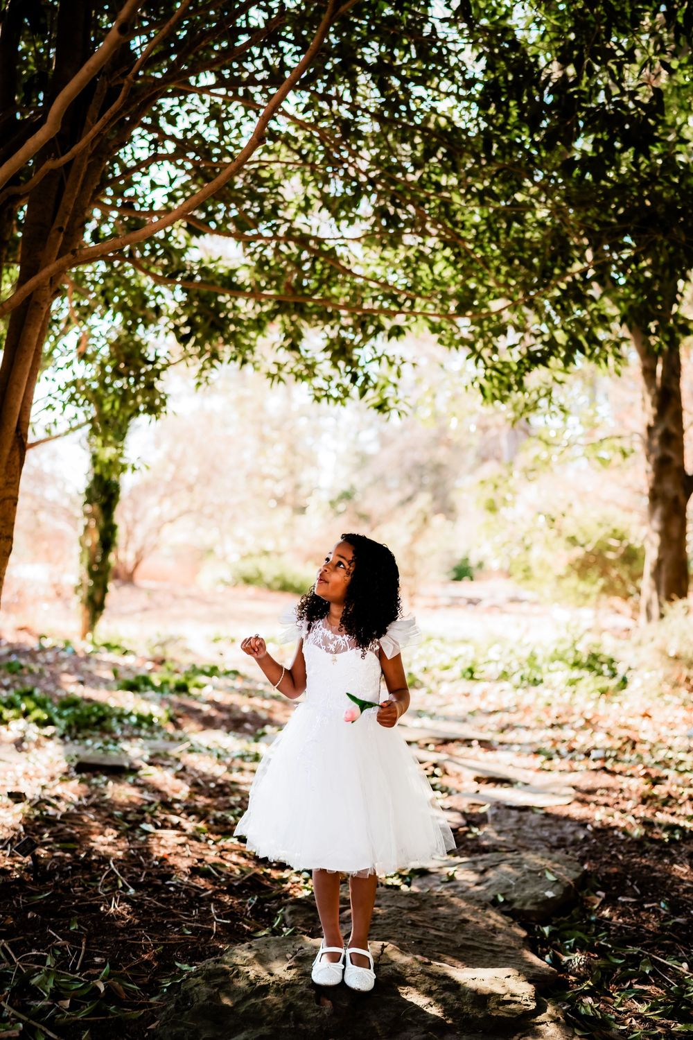 Young Black girl in white dress standing in sunlit park, looking up at trees, wearing white shoes.