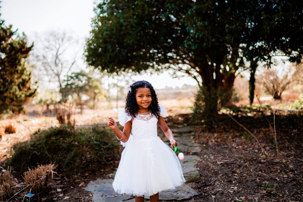 Young girl in white flower girl dress smiling outdoors among trees and natural landscape.