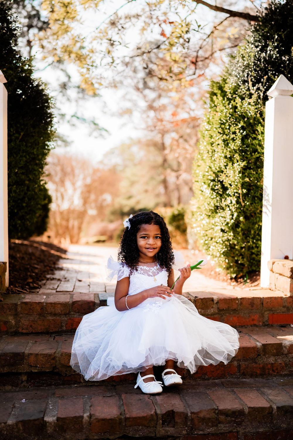 Young girl in white lace tutu dress sitting on brick steps in a garden.