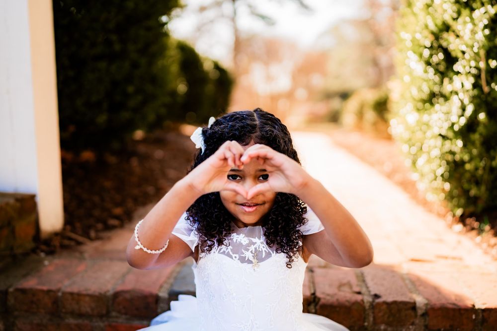 Young girl in white dress making heart shape with hands outdoors.