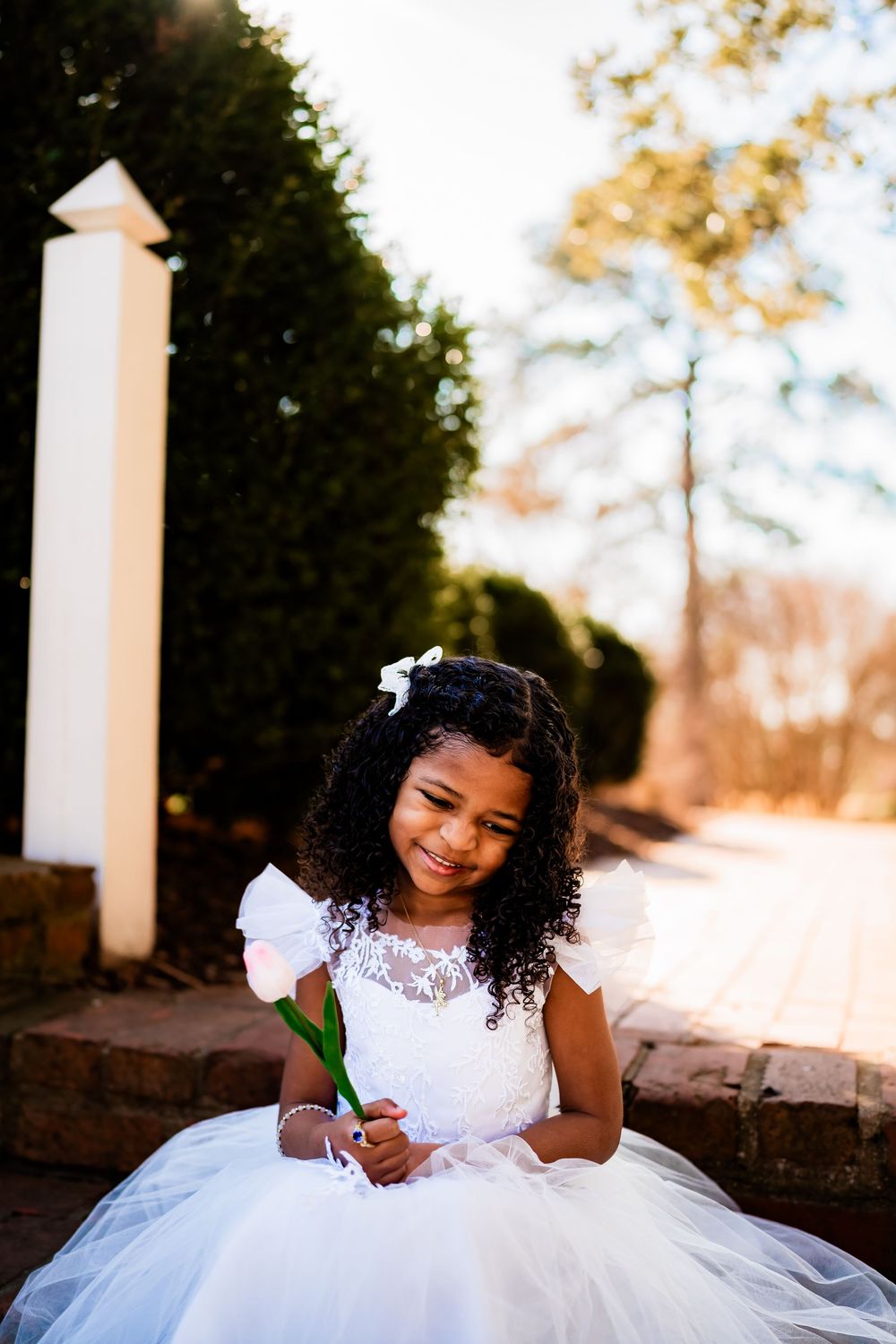 Young flower girl in white tulle dress holding pink tulip, smiling outdoors at wedding venue.