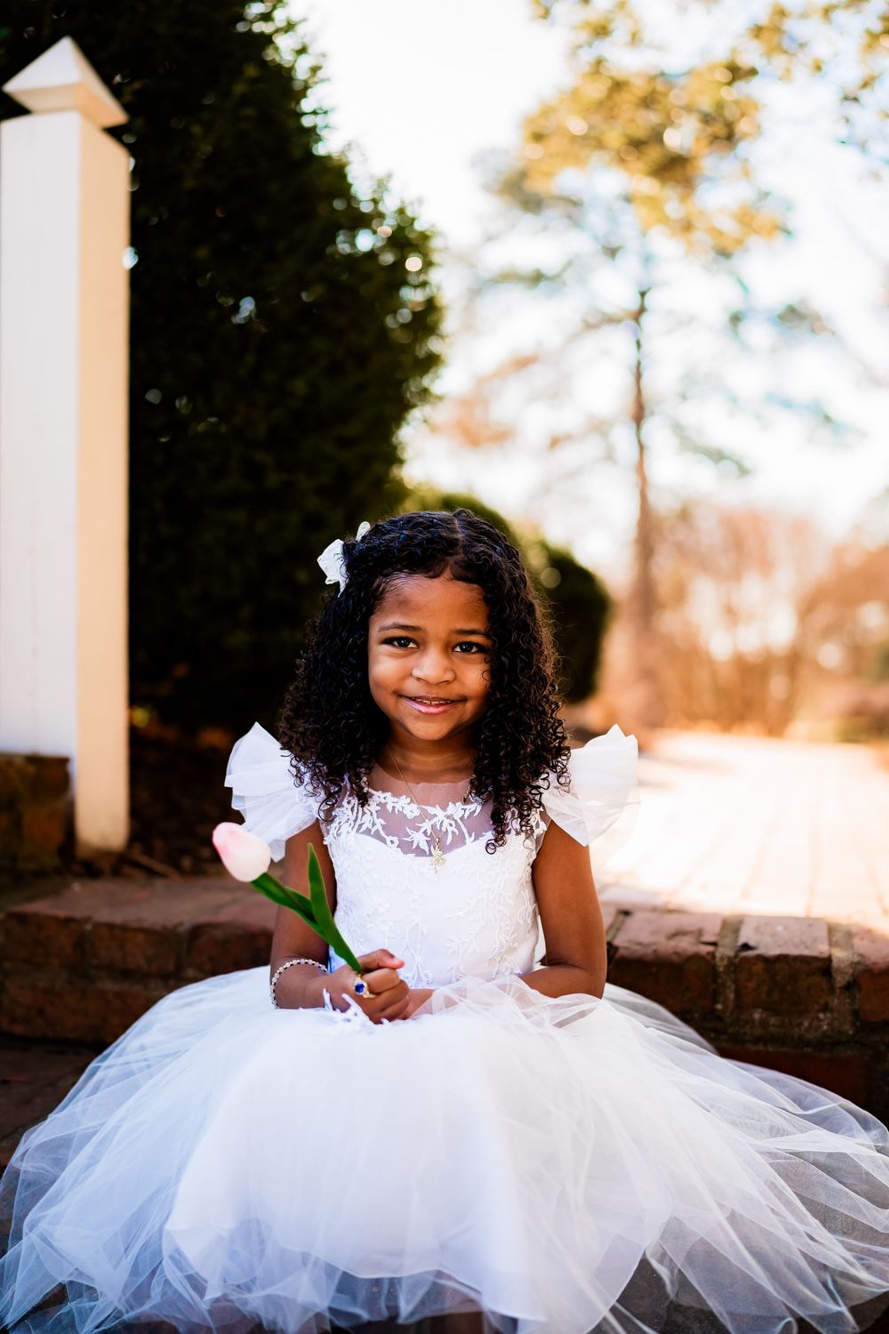 Smiling young girl in white lace dress holding a pink flower outdoors.