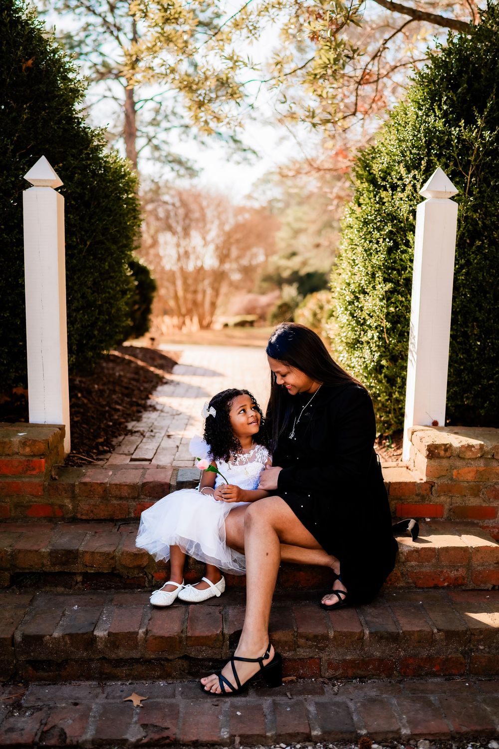 A woman in black dress sits on brick steps with a little girl in white dress in a garden pathway.