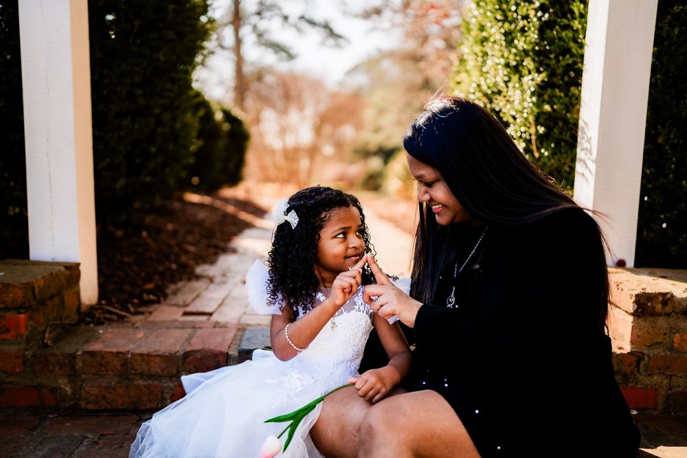 Mother and young daughter in white dress sharing a tender moment on garden pathway with white flower.