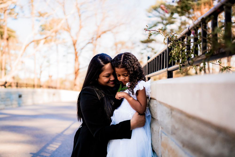 A mother in black embraces her young daughter in a white dress outdoors near a stone wall and pond.