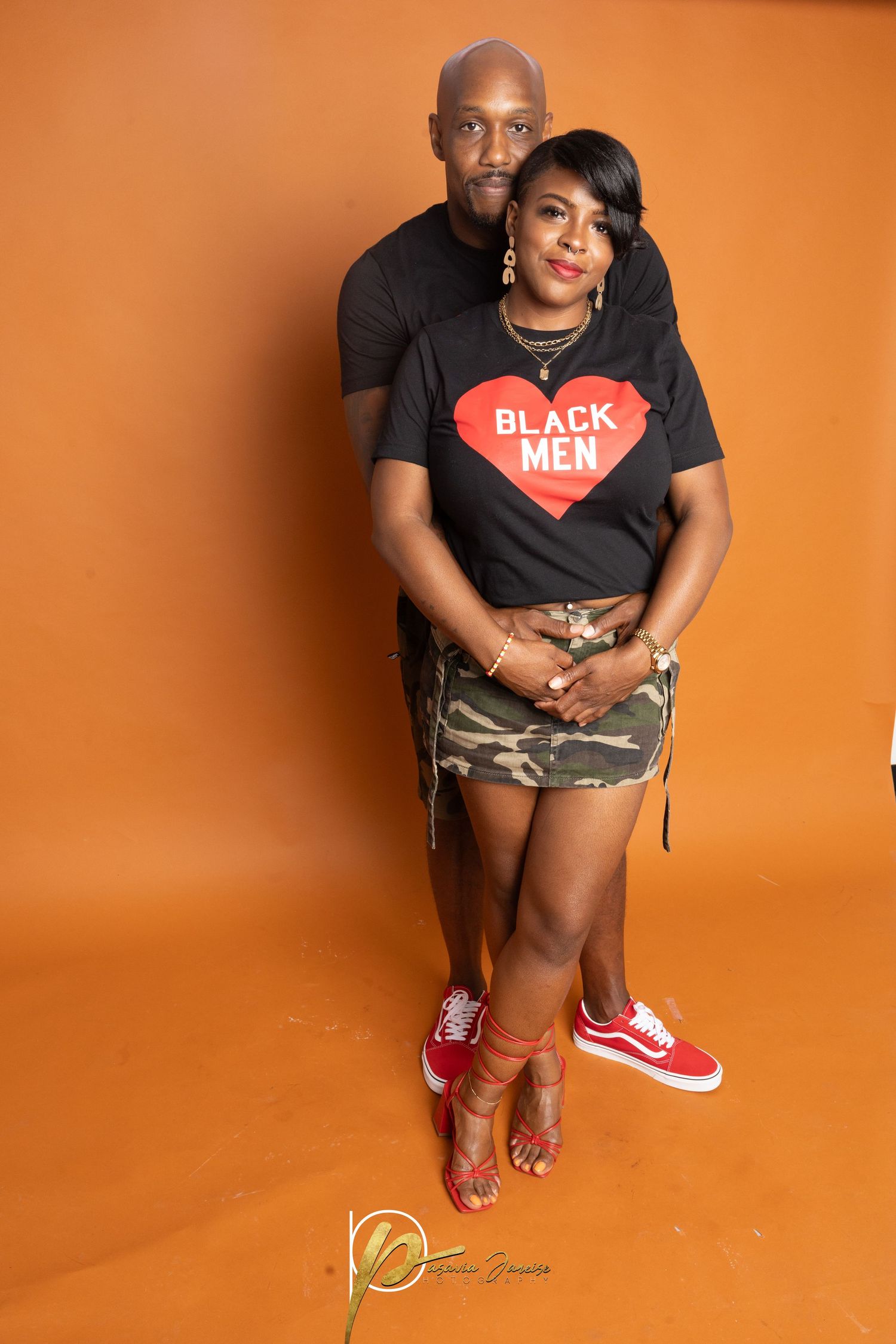 Couple posing together against orange background, woman wearing Black Men t-shirt, camo skirt, and red Vans sneakers.