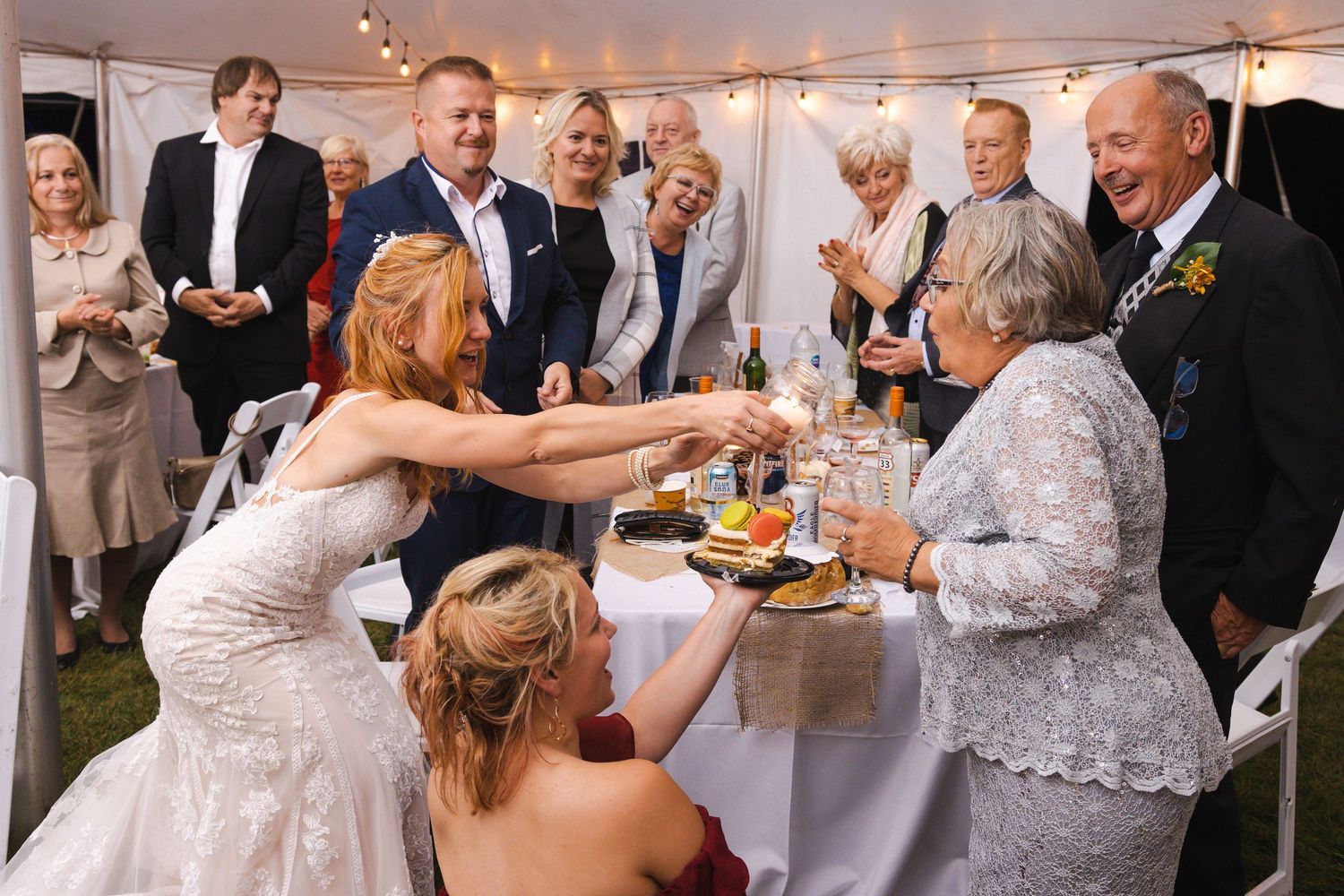 Wedding guests gather around a table under a white tent during a reception celebration.