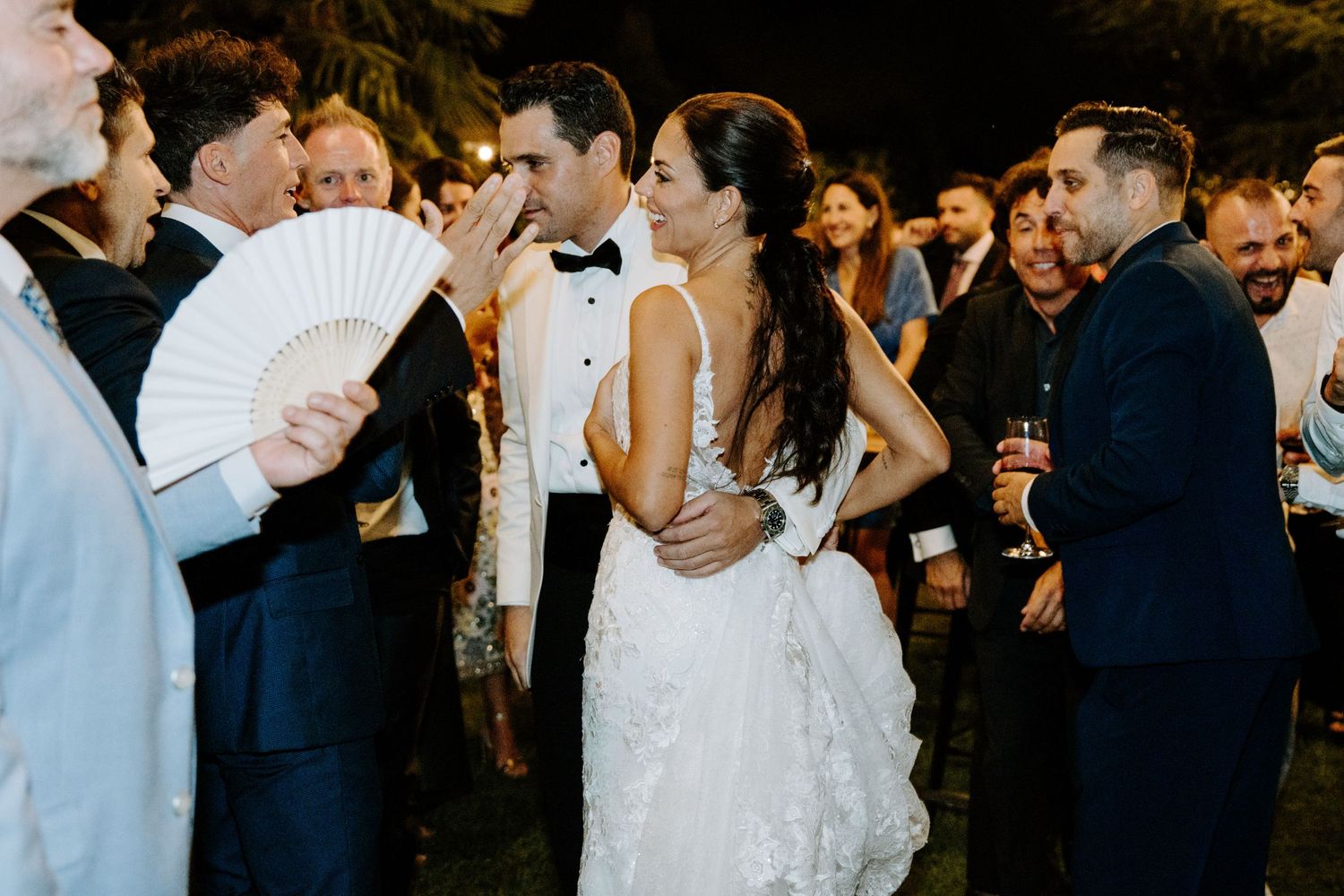Bride and groom surrounded by cheering wedding guests during outdoor evening reception celebration.