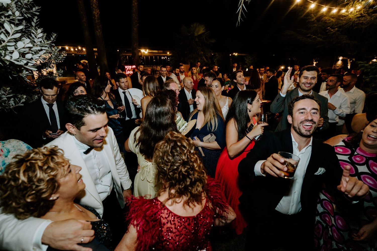 Joyful wedding guests dancing and celebrating at an outdoor evening reception under string lights.