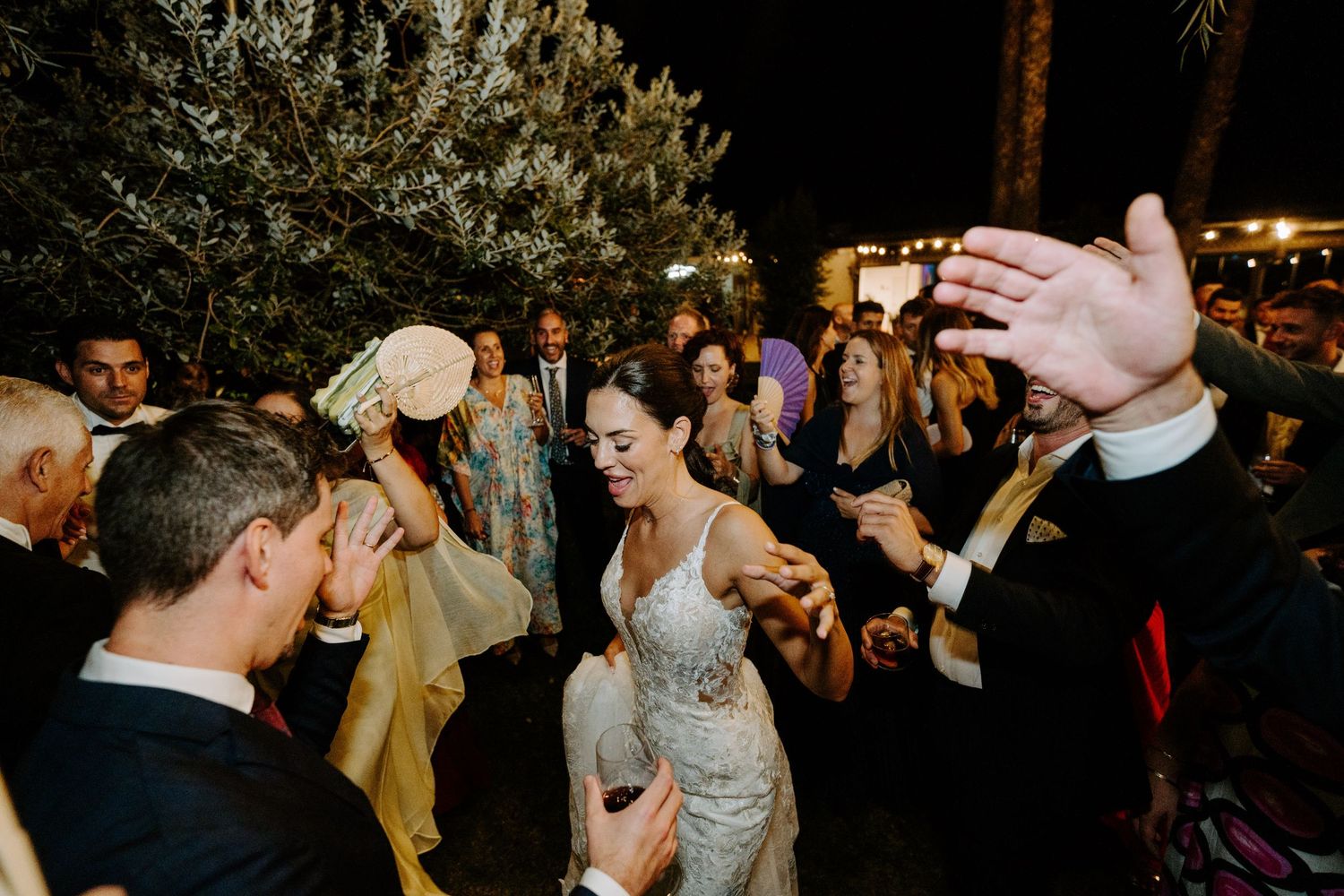 Bride dancing joyfully with guests at an outdoor wedding reception at night.