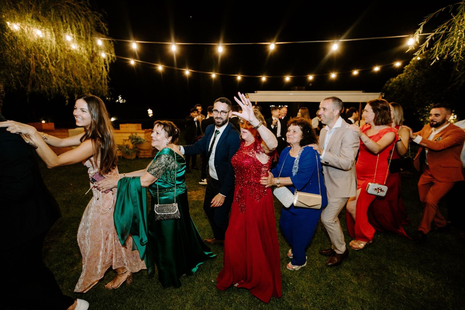 Wedding guests dance joyfully under string lights at an outdoor evening reception celebration.