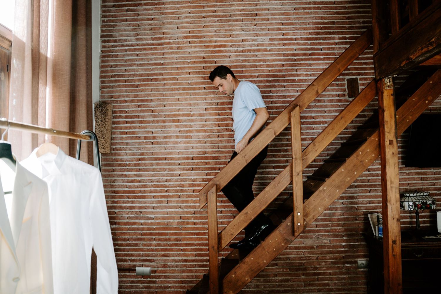 Man in white shirt walks down rustic wooden staircase in brick-walled room with hanging clothes.