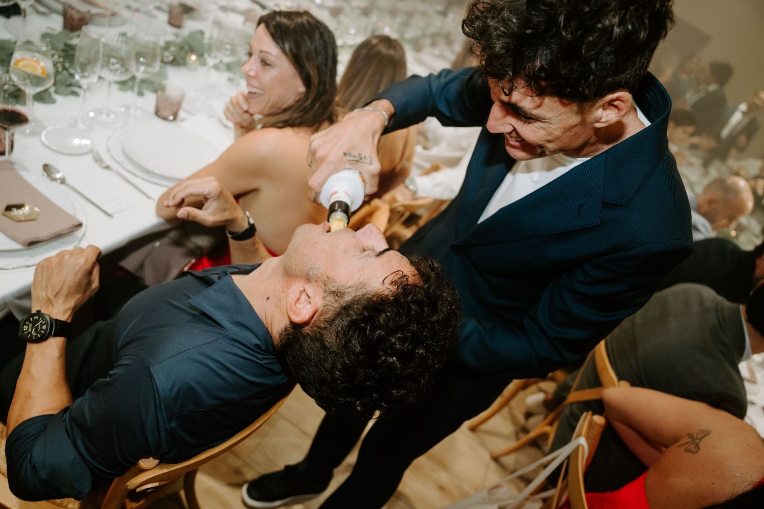 Groom pours champagne into best man's mouth at wedding reception dinner table.