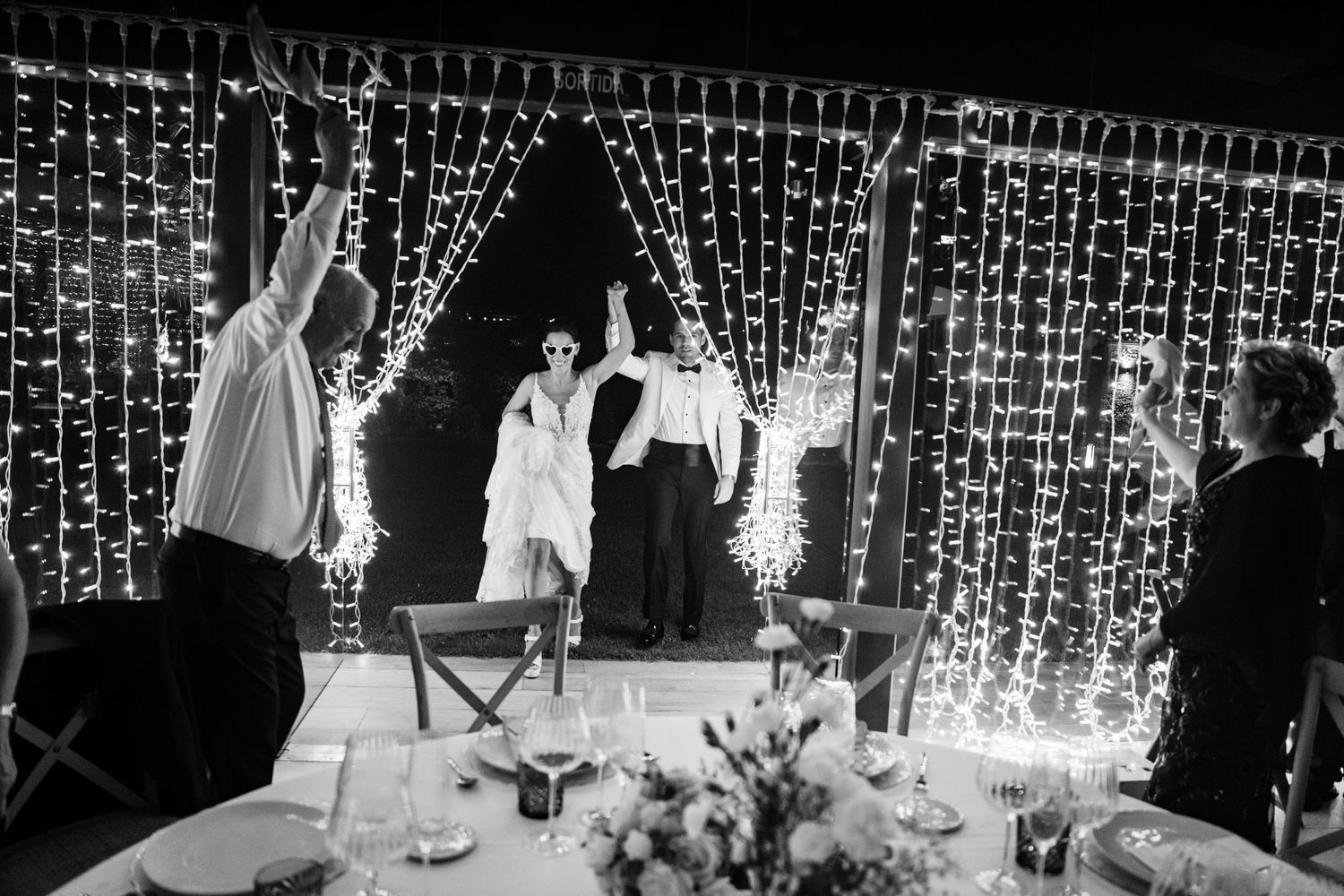 Black and white photo of newlyweds celebrating joyfully in front of sparkling fairy light backdrop.