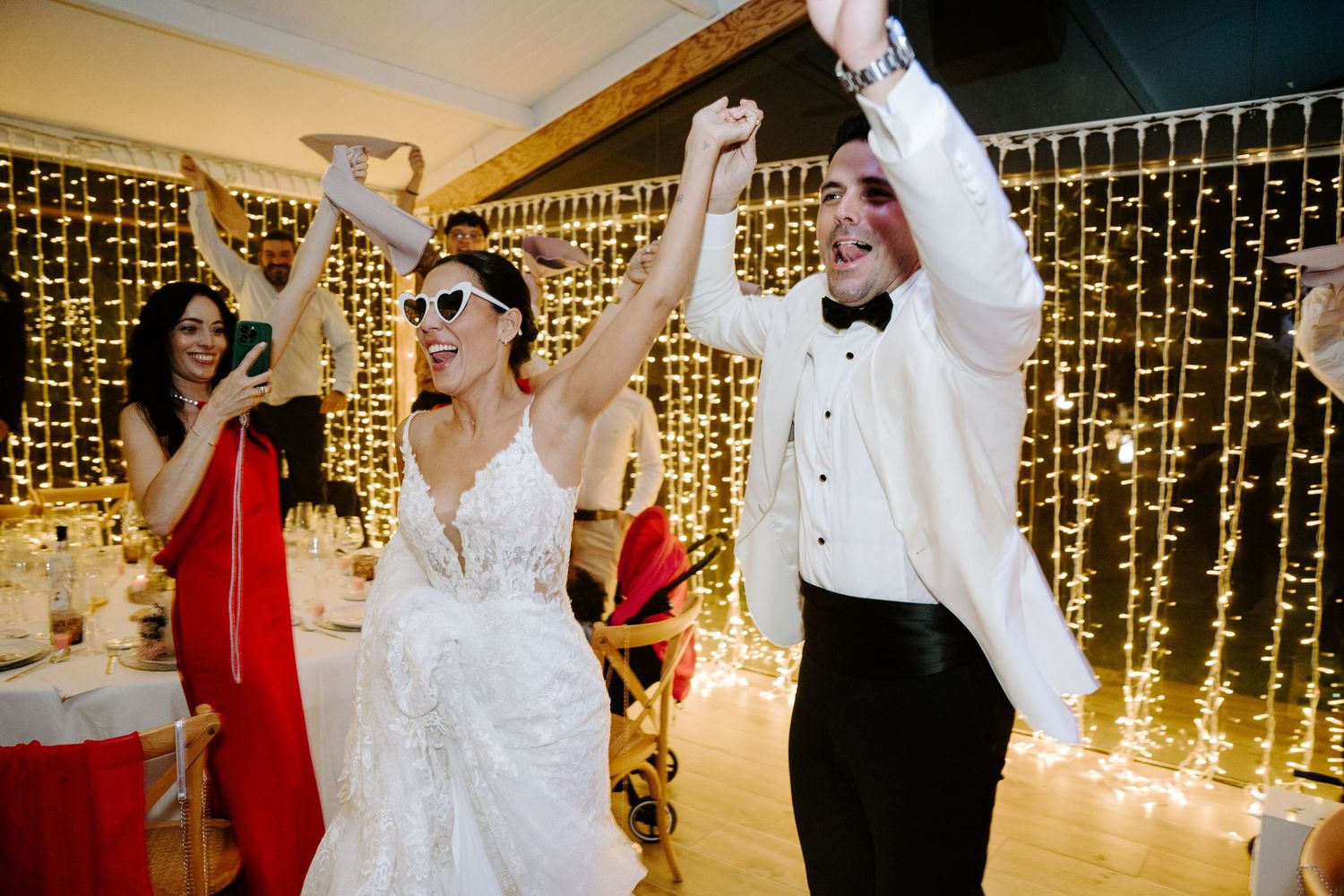 Joyful bride and groom celebrating with raised hands under warm fairy light curtain.
