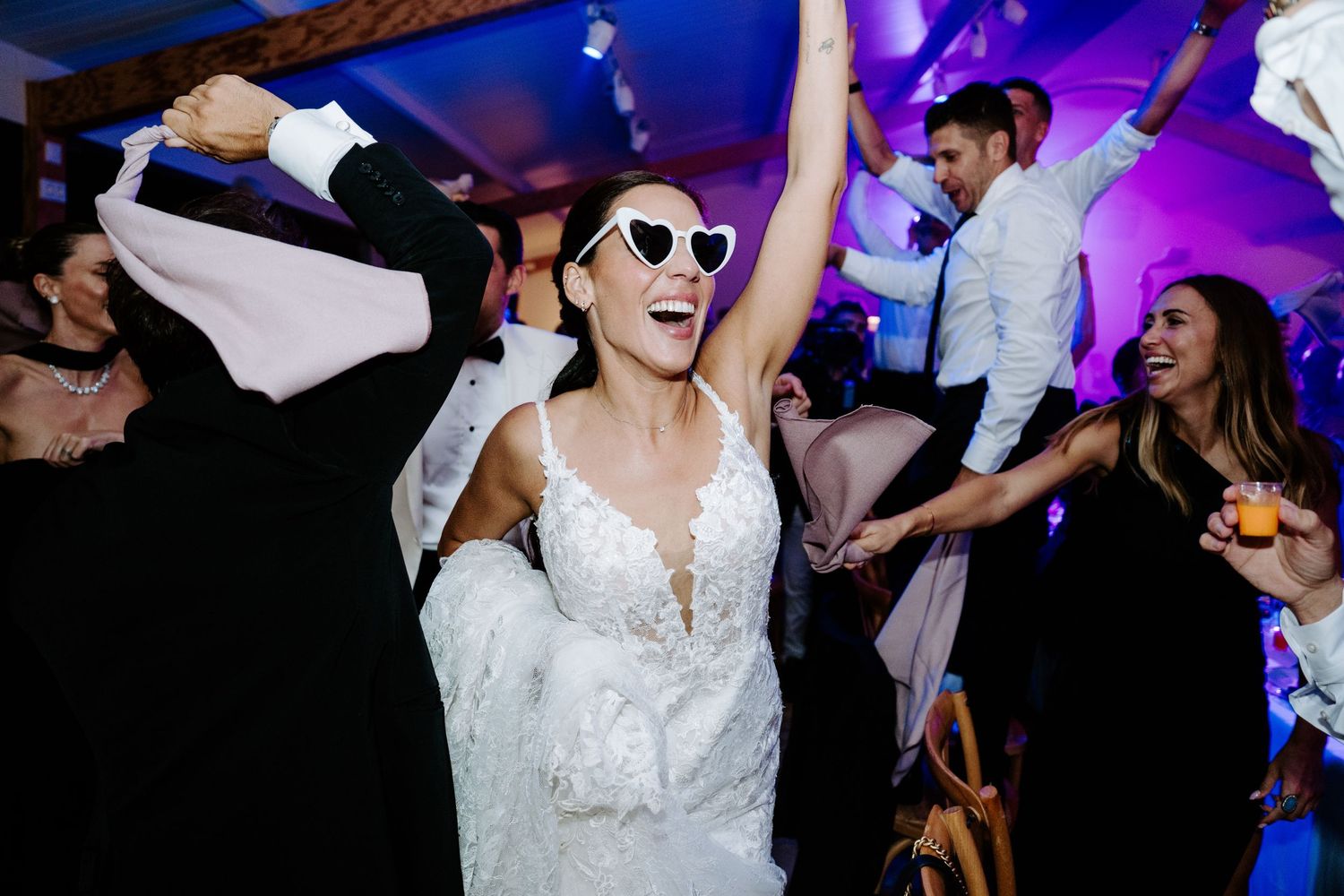Joyful bride in white gown wearing heart-shaped sunglasses dancing at wedding reception.