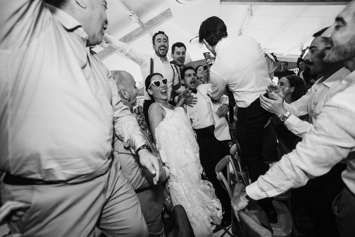 Black and white photo of lively wedding reception with guests celebrating and groom lifted by crowd.