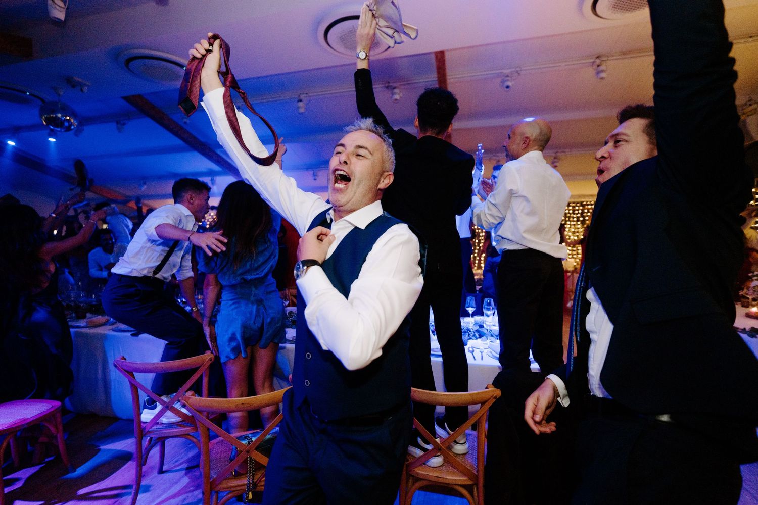 Man in vest celebrates at wedding reception, holding tie overhead while dancing with guests under blue lights.