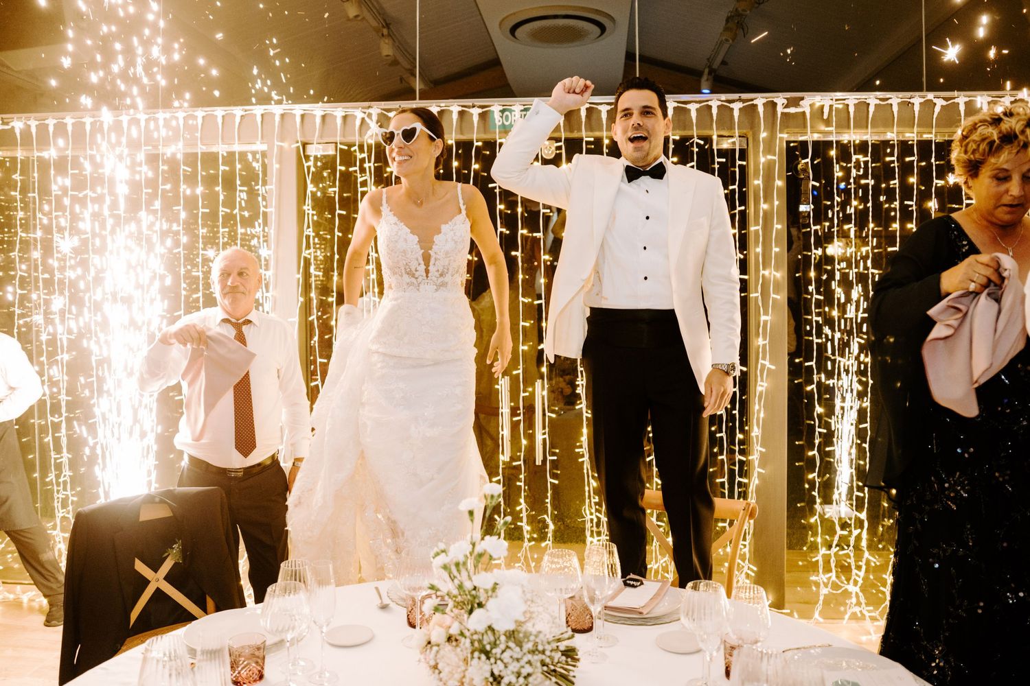 Bride and groom celebrate with sparklers and string lights during dramatic wedding reception entrance.