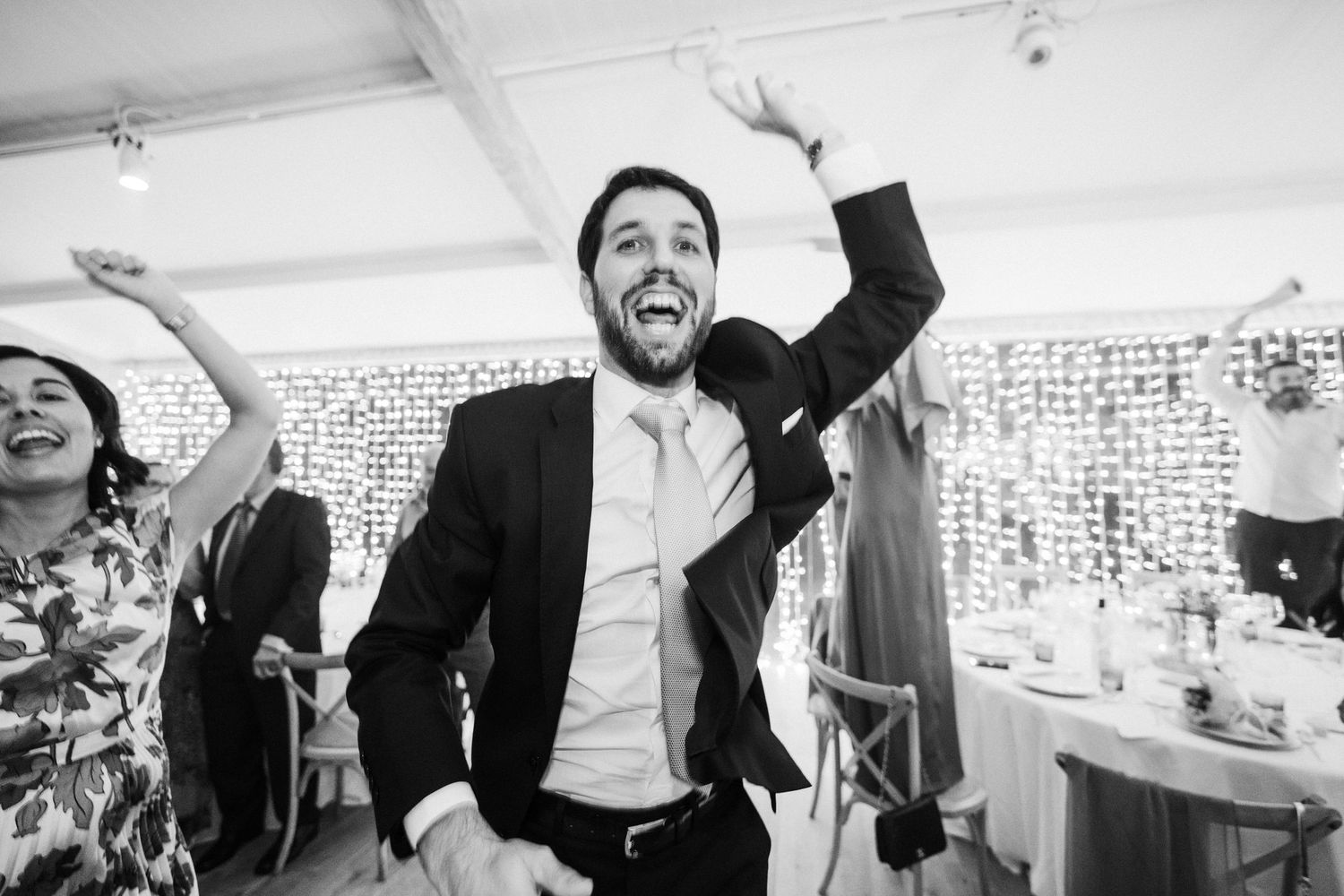 Joyful man in black suit dances with arms raised at a festive wedding reception.