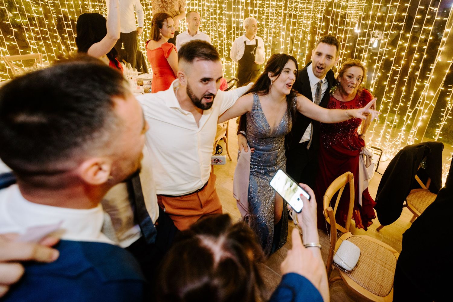 Lively wedding reception guests dancing and celebrating in front of glowing string light backdrop.