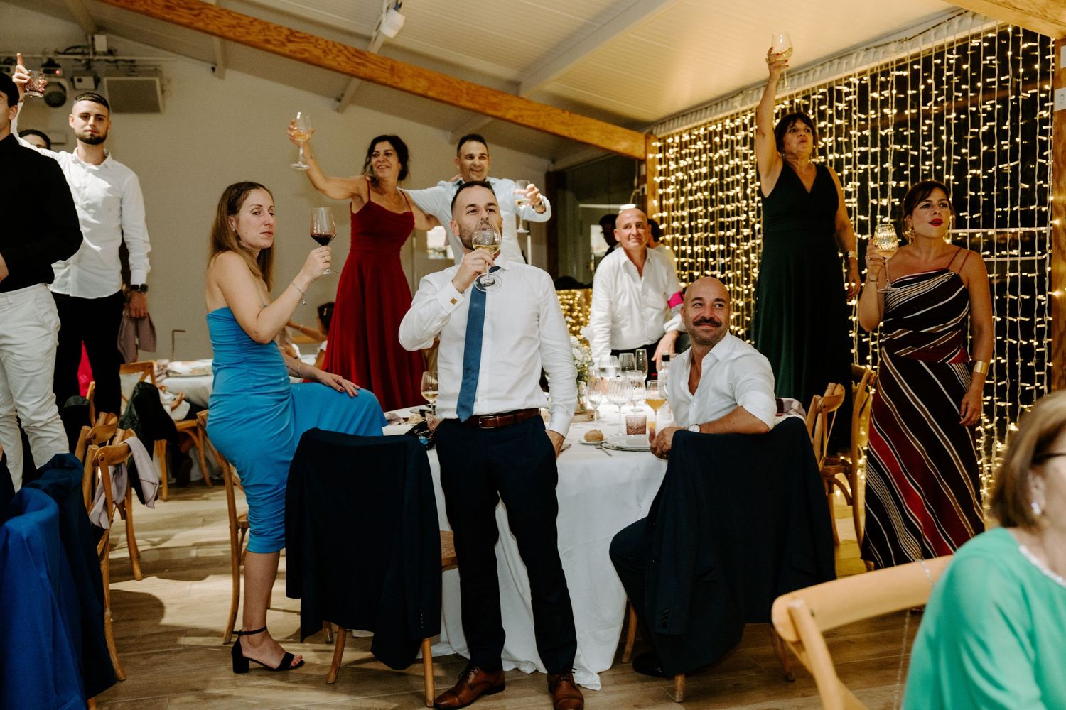 Wedding guests raising glasses in a toast during indoor reception with fairy light backdrop.