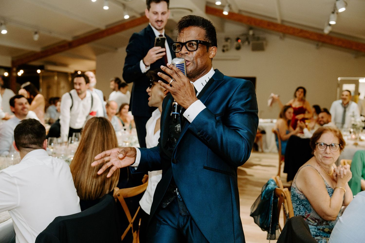 Charismatic wedding MC in navy blue suit delivering speech to guests at indoor reception venue.