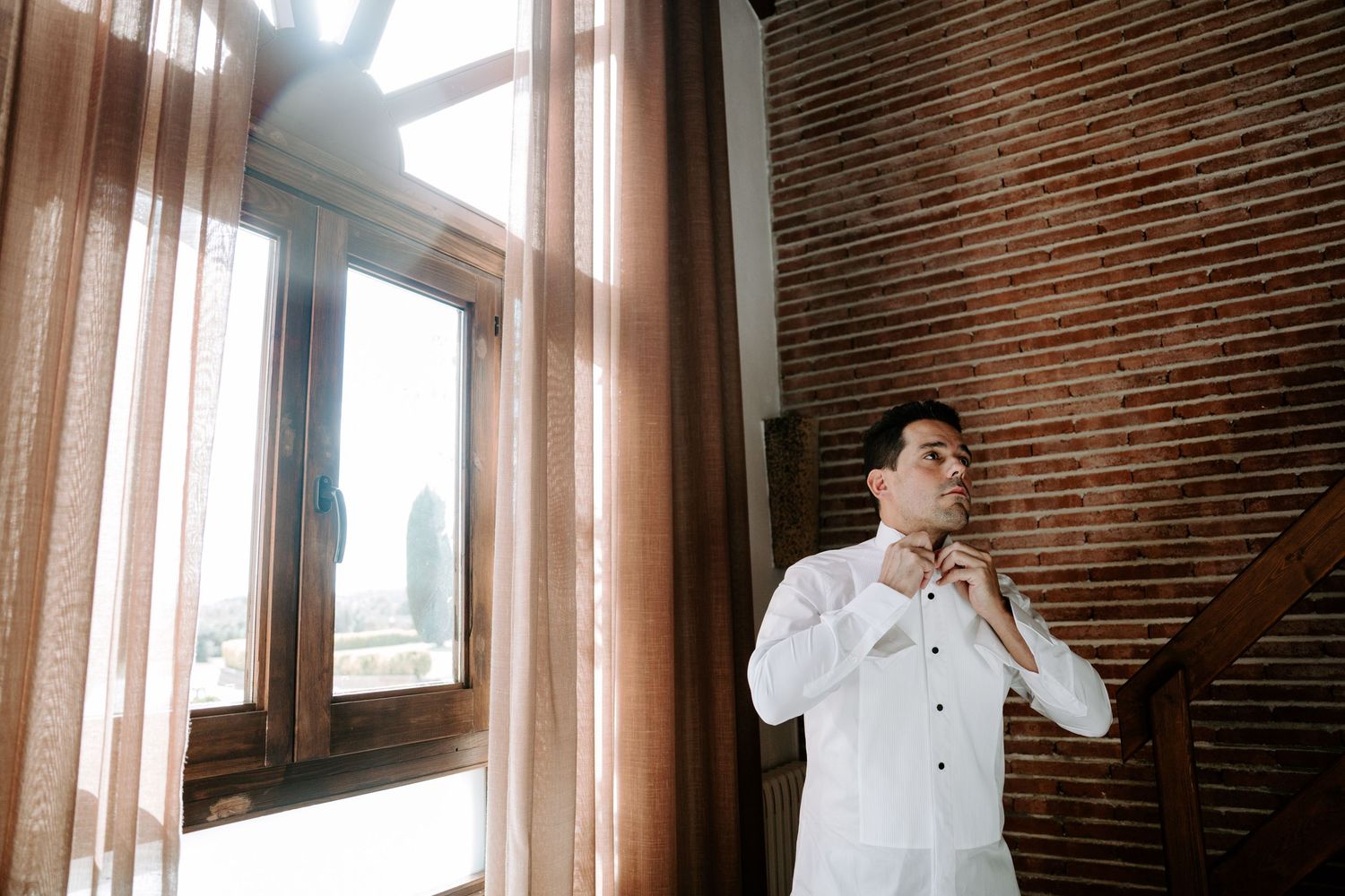Groom adjusting collar in white shirt standing by sunlit window on wedding day.