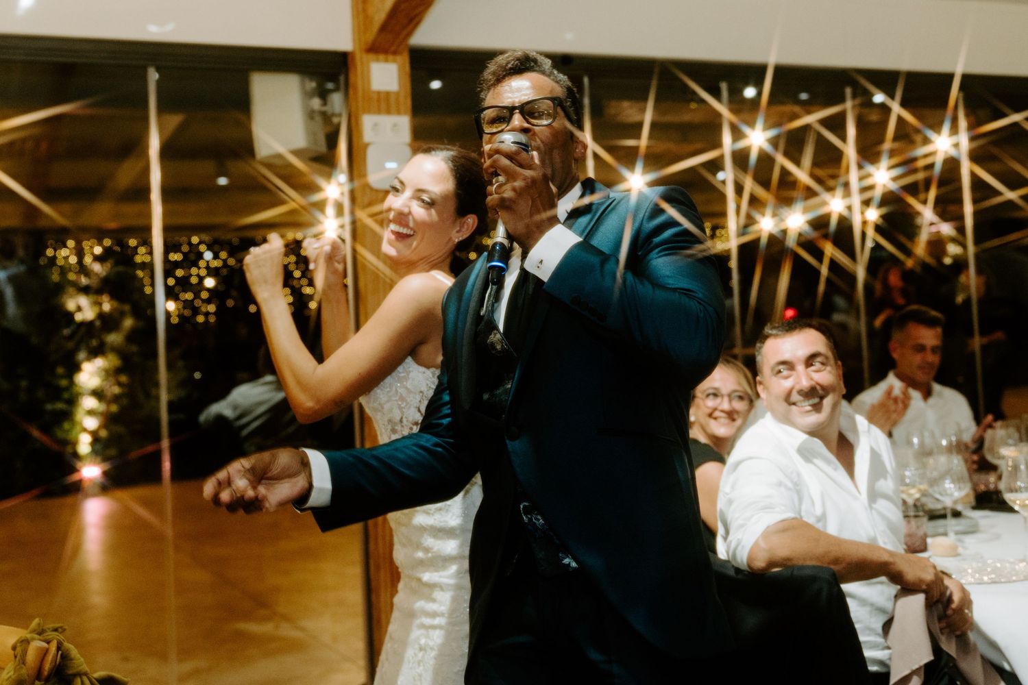 Bride and groom dancing joyfully at wedding reception with guests smiling in background.
