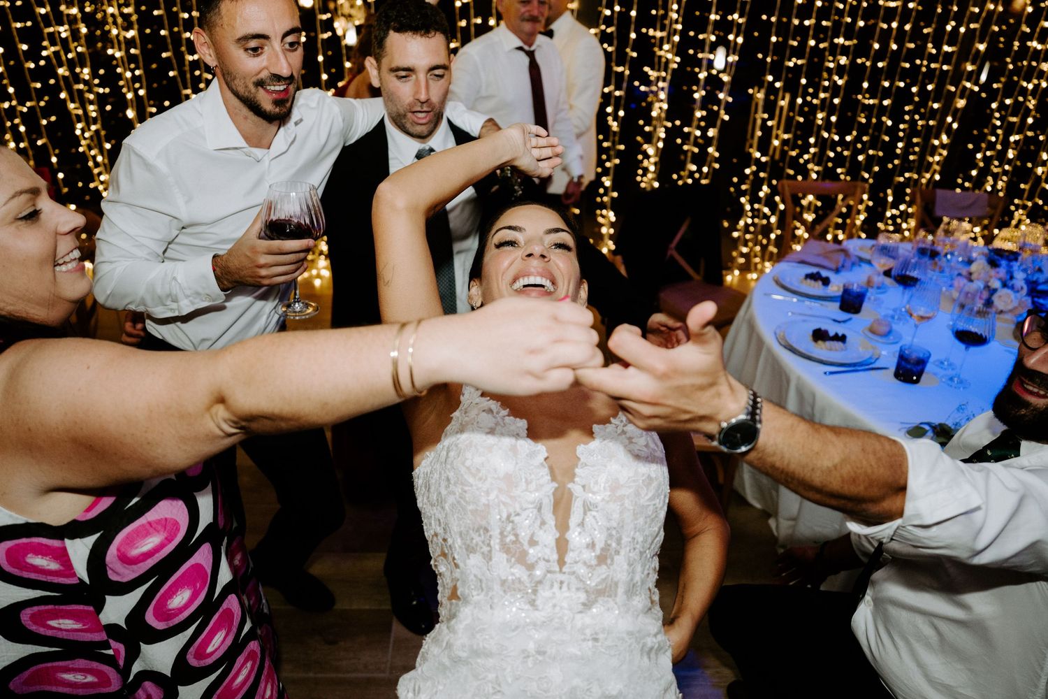 Bride laughs joyfully as guests dance around her at wedding reception with warm fairy light backdrop.