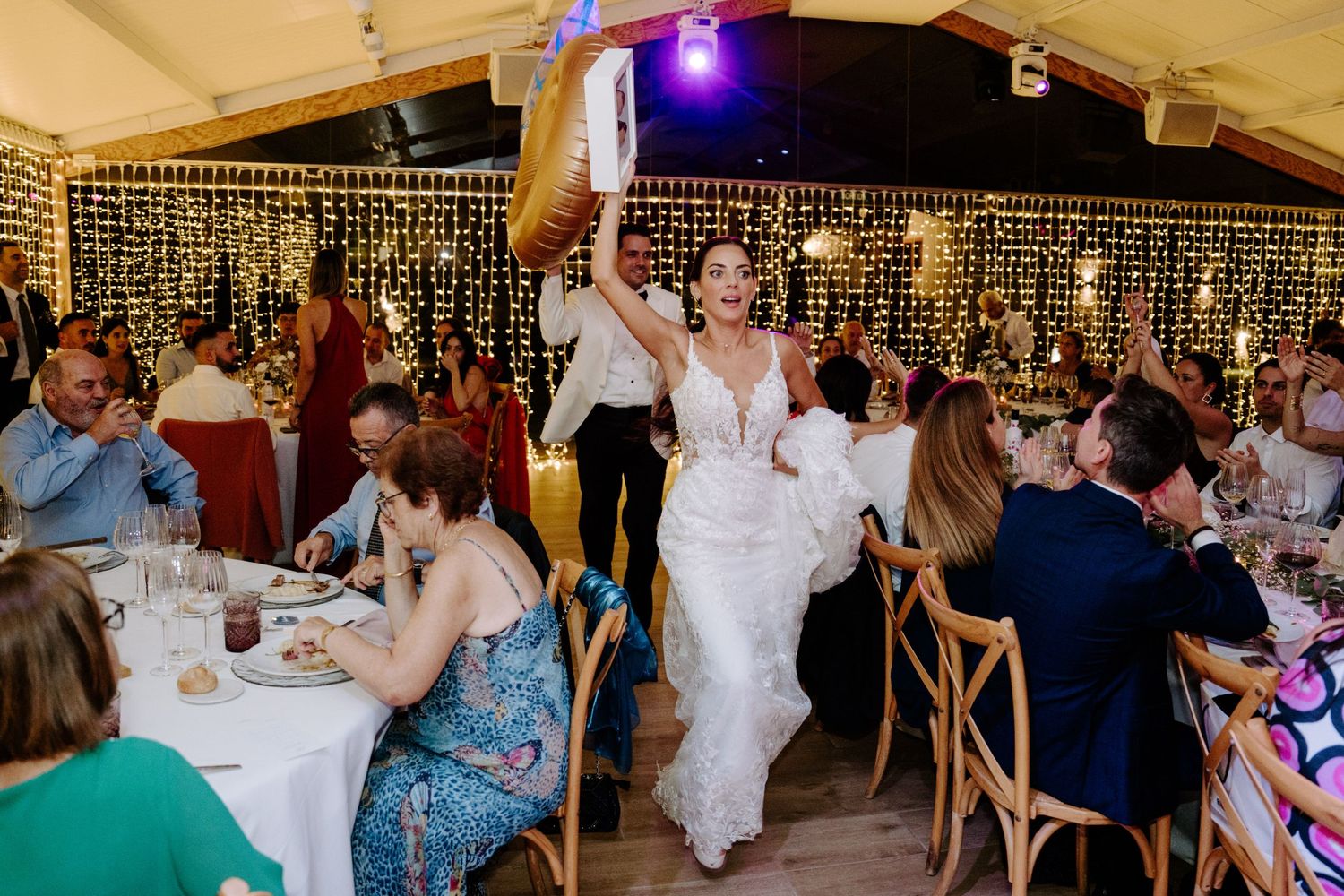 Bride in lace gown walks triumphantly through wedding reception holding item above her head.