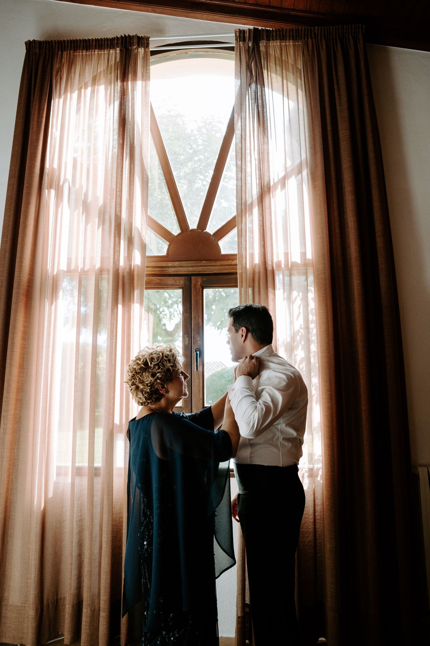 Groom being helped with his tie by a woman near a sunburst window with flowing curtains.