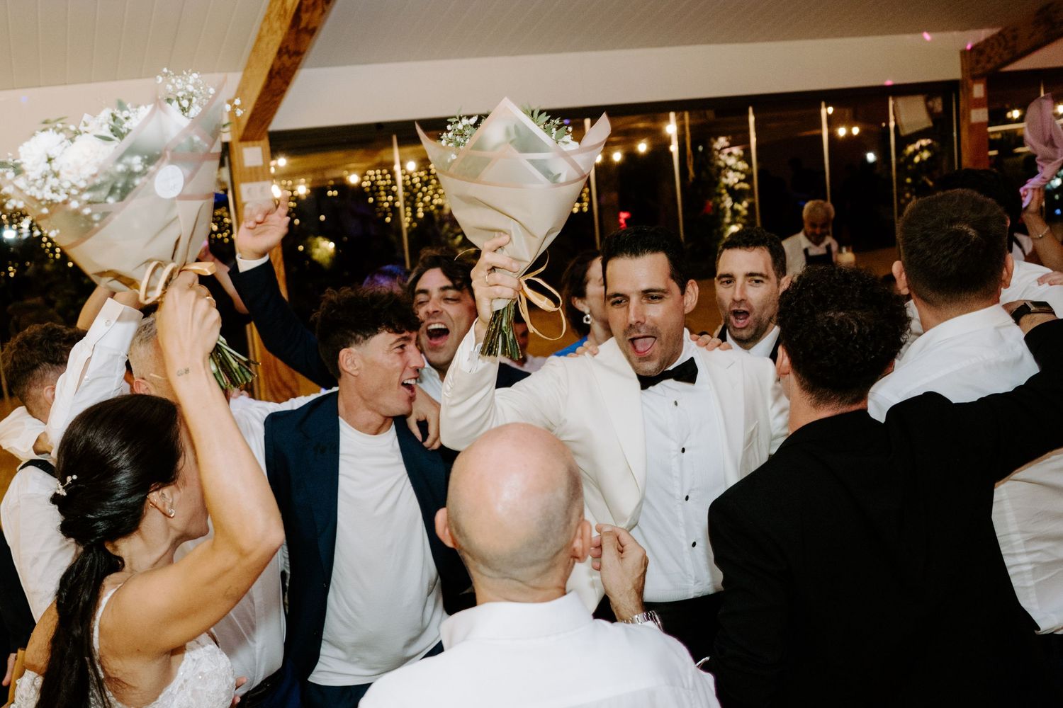 Groom in white tuxedo celebrated by cheering wedding guests holding floral bouquets indoors.