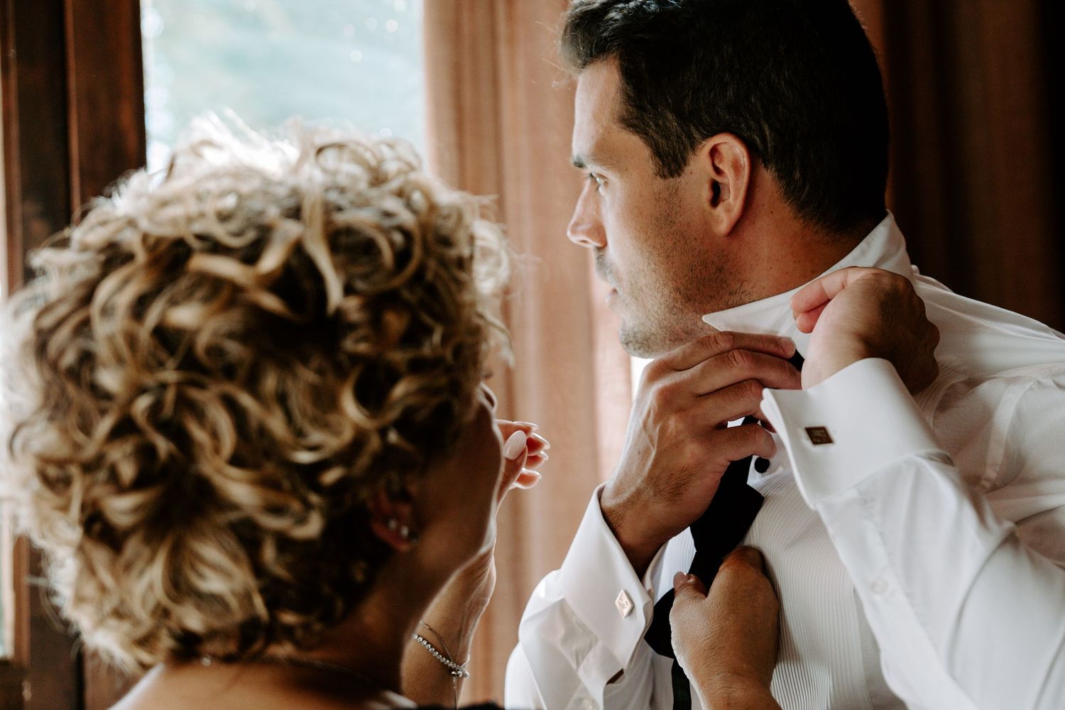 Woman helps groom adjust tie and collar before wedding ceremony indoors.