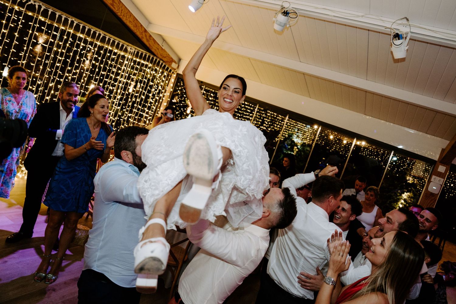 Bride in white gown being lifted by guests during energetic wedding reception celebration.