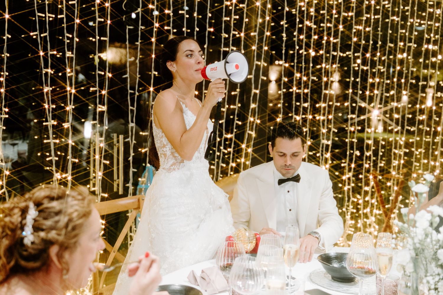 Bride in white gown speaks into megaphone at wedding reception table with fairy light backdrop.