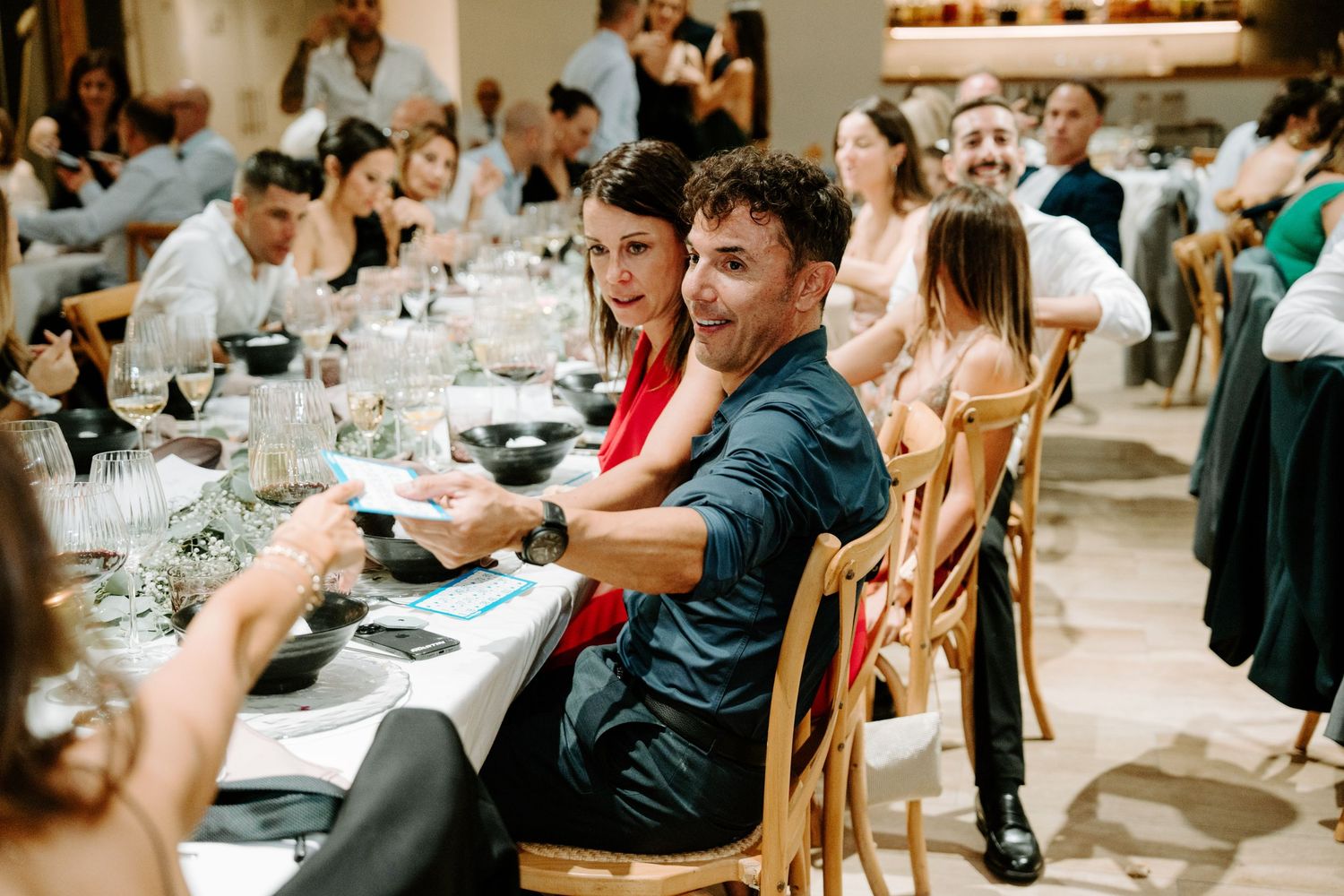 Excited wedding guests laugh and react at elegantly set dinner table during indoor wedding reception.