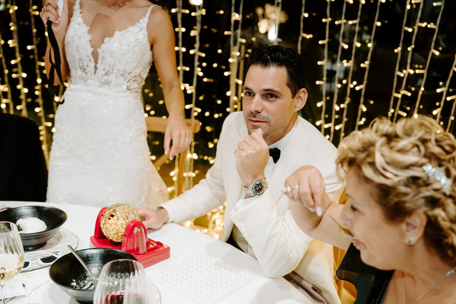 Groom in white tuxedo sits at wedding reception table with fairy lights backdrop.