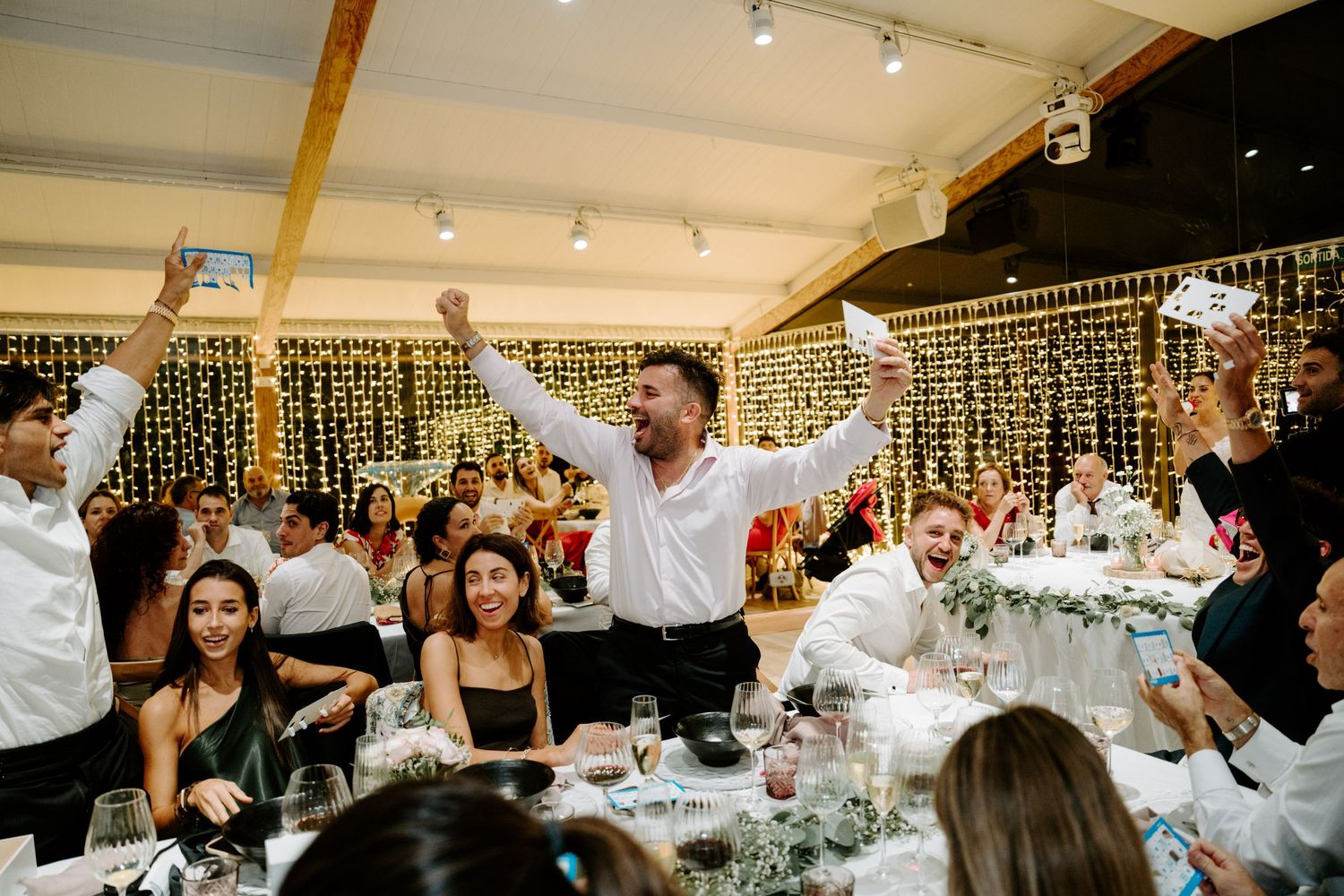 Excited wedding guests celebrating with arms raised at elegant reception with fairy light backdrop.