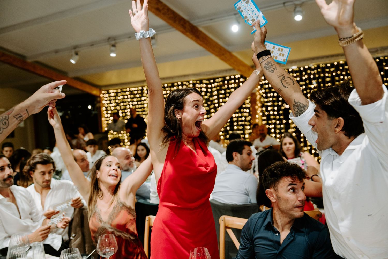 Excited guests celebrating at a wedding reception with fairy lights, raising hands and cheering joyfully.